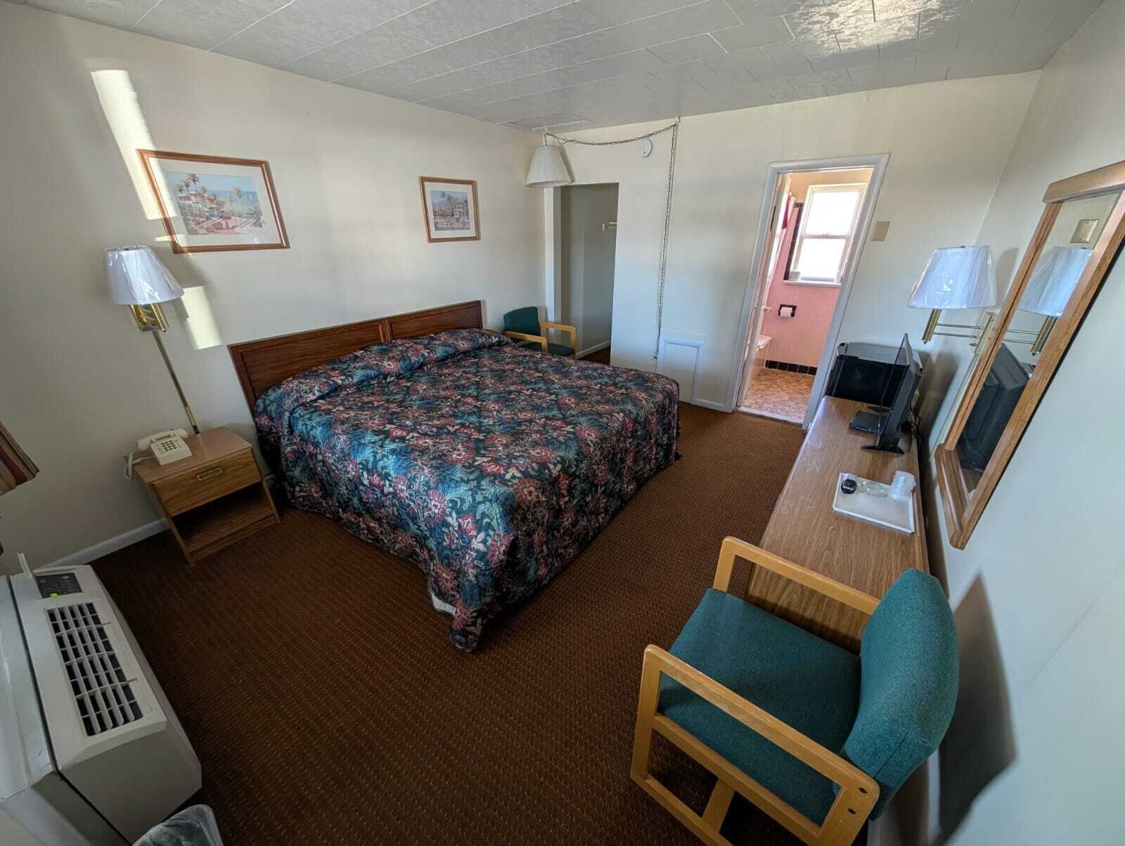 A hotel room with a bed, a desk, a chair, a lamp, and a TV.