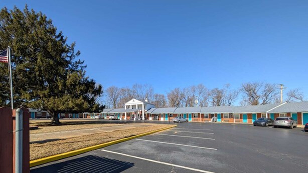 A motel with a central building and several attached units, a parking lot, and a flagpole. - Relax Inn (New Castle)