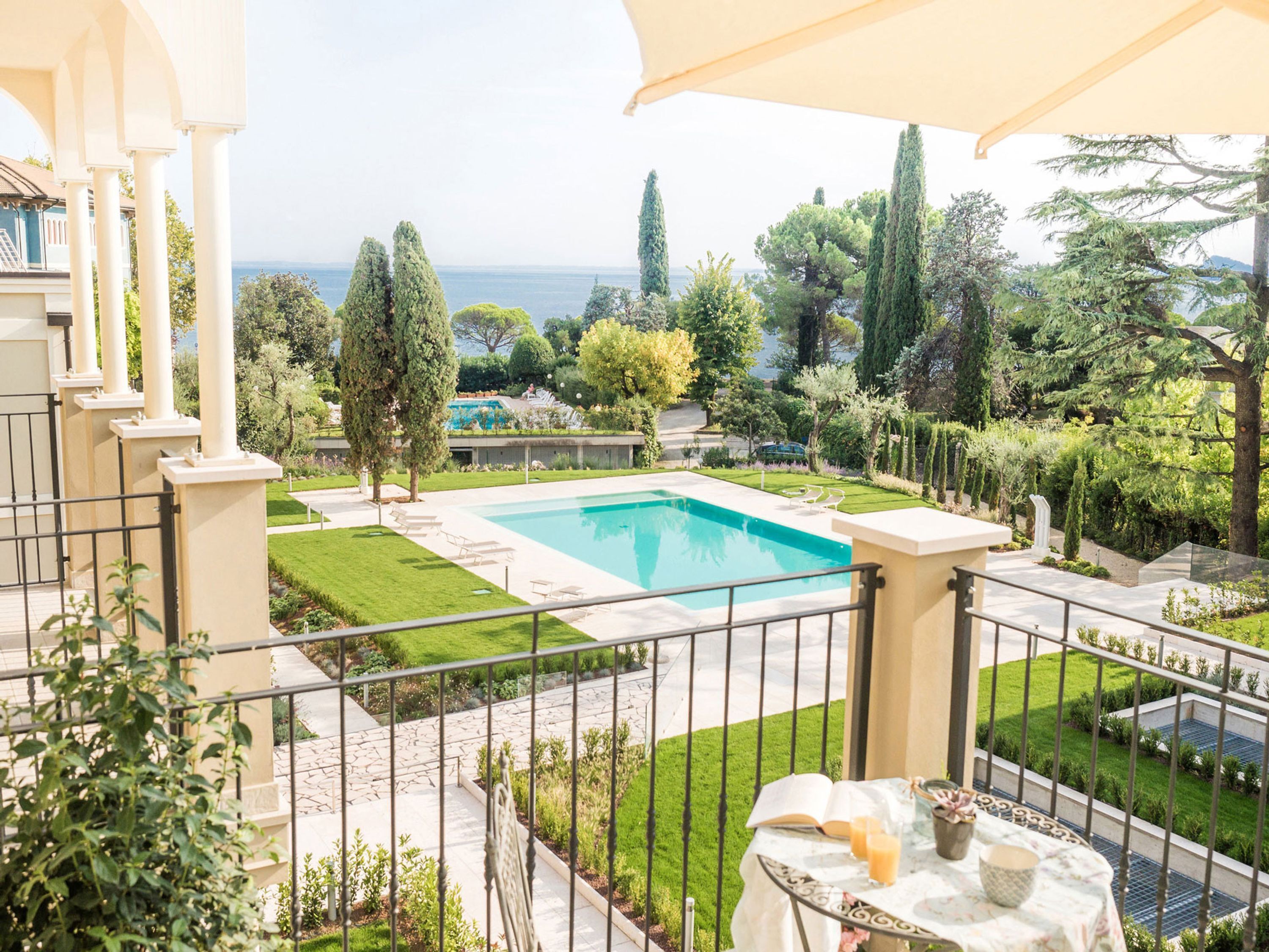 A pool area with a view of the sea, surrounded by greenery and a white fence.