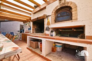 Une cuisine extérieure dotée d’un four en briques, de poutres en bois et d’un coin repas avec une table dressée pour deux personnes.