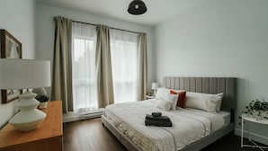 Un dormitorio moderno con una cama grande, una mesita de noche con lámpara, una pequeña mesa auxiliar con una planta y una ventana con cortinas.