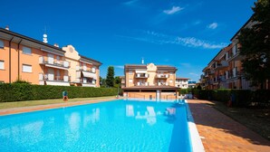 A swimming pool surrounded by apartment buildings with balconies, a clear blue sky, and a hedge along the poolside. - Appartamento Francesca (Sirmione)