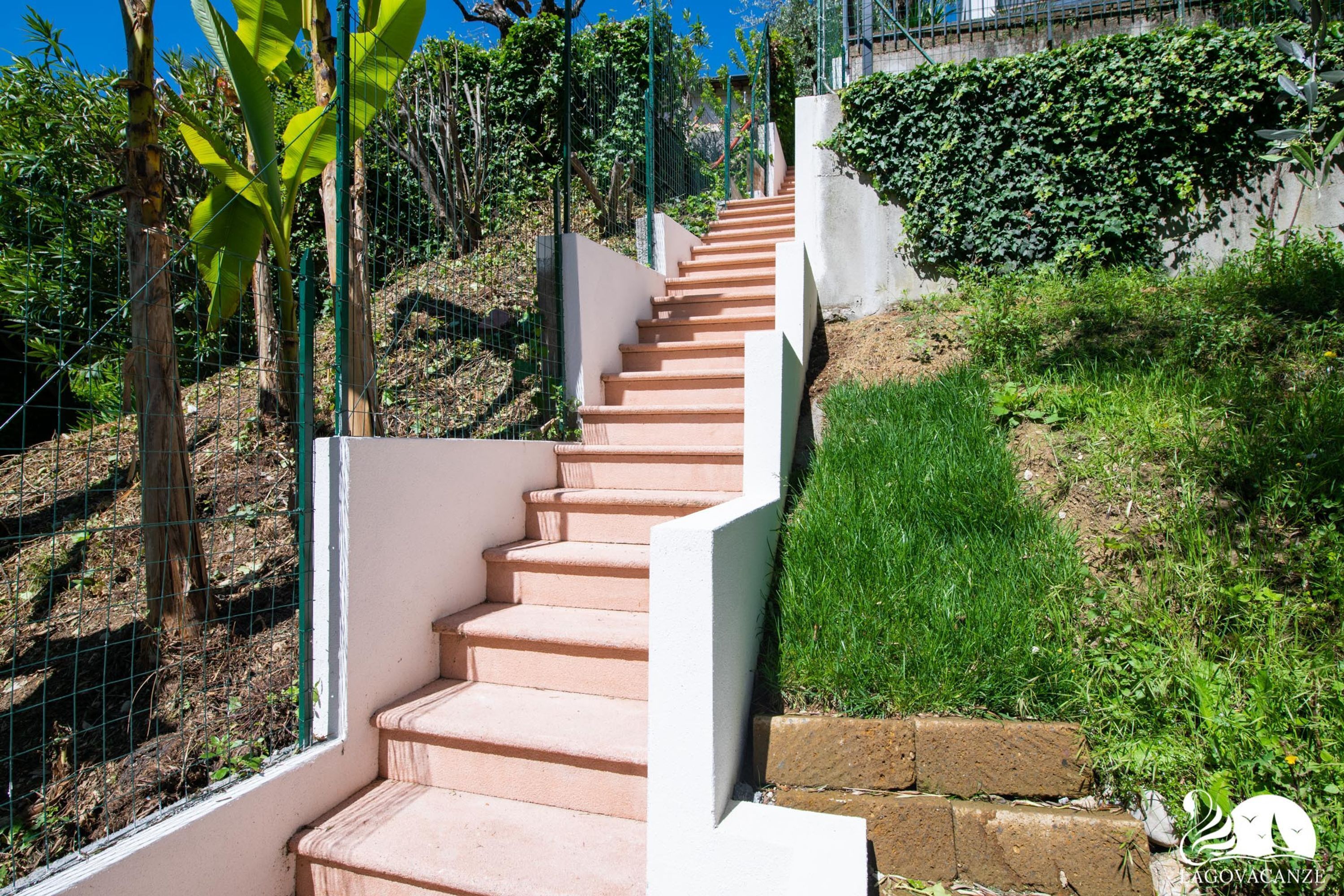 A staircase with a concrete wall on one side and a metal fence on the other, surrounded by greenery and a clear blue sky.