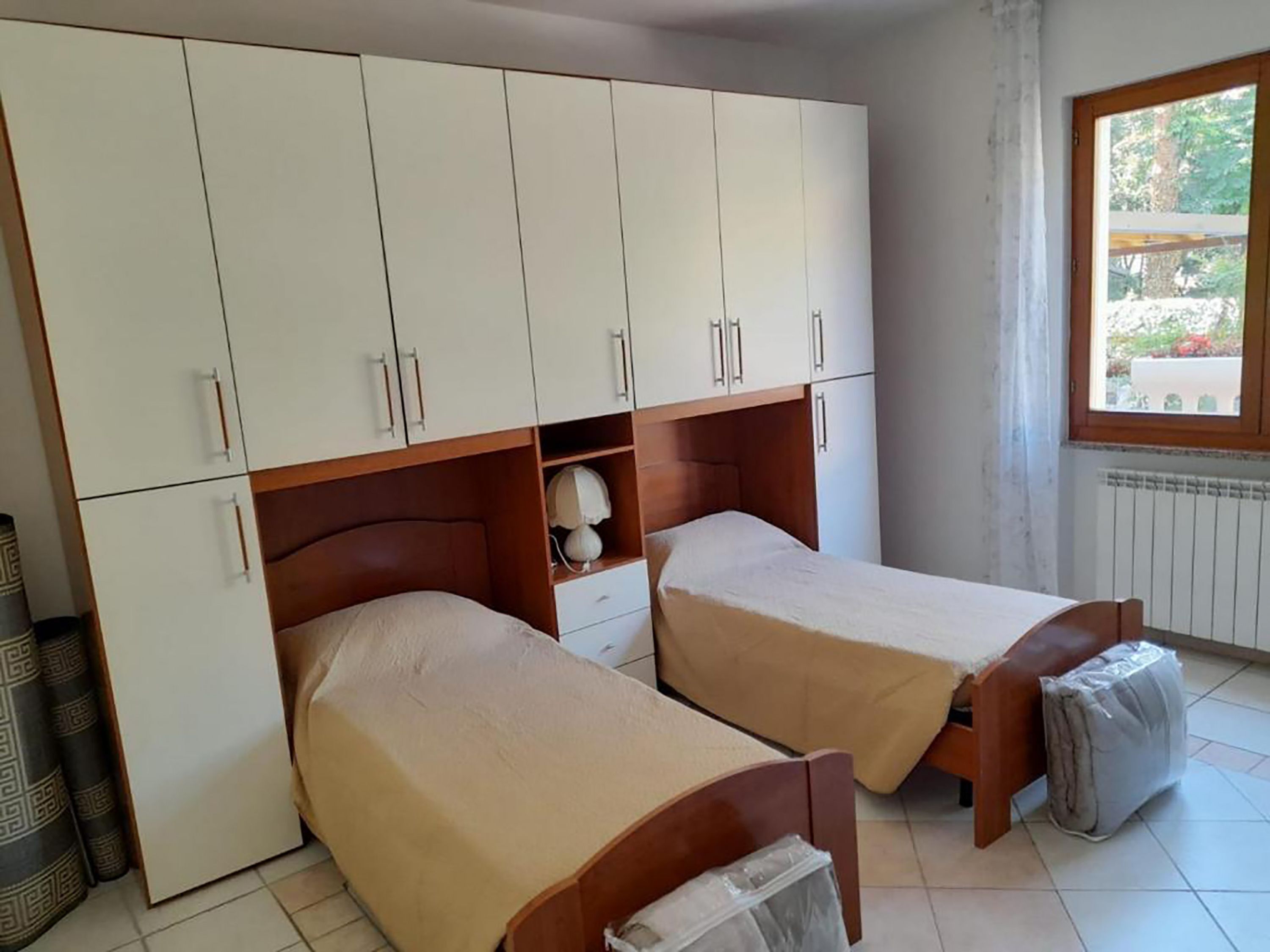 A room with two single beds, a wardrobe, a small table, and a window with a view of greenery.