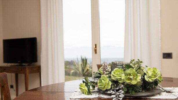 A dining table with a floral centerpiece, a television on a stand, and a view of the outdoors through a door. - Villa Jole in Polpenazze del Garda (Polpenazze del Garda)