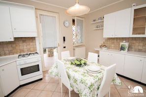 Une cuisine avec des meubles blancs, une cuisinière à gaz, une table à manger recouverte d’une nappe à motifs floraux et des chaises.