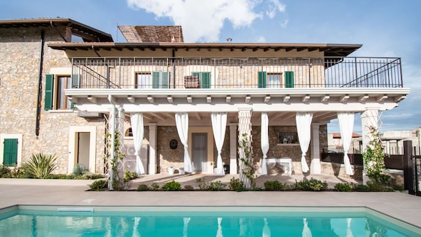Una casa de piedra de dos pisos con balcón, piscina y zona de estar exterior cubierta.