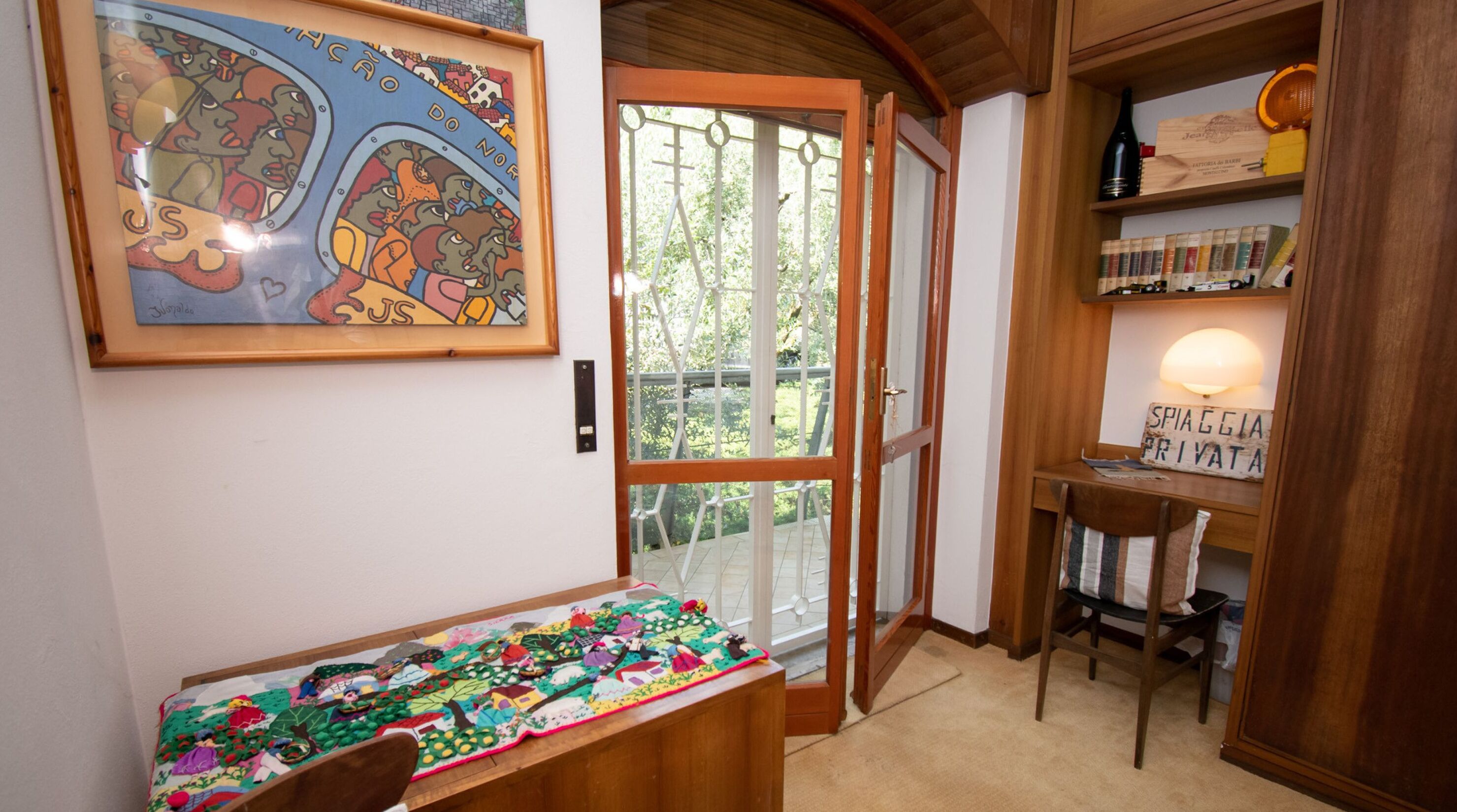 Een kamer met een houten deur, een lijst met een kunstwerk, een tafel met een kleurrijke quilt en een plank met boeken en een fles.