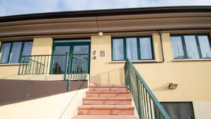 Un edificio con fachada color beige, marcos de ventanas verdes y una escalera con barandilla verde que conduce a una puerta.