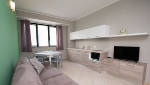 Una sala de estar moderna con un sofá color beige, una mesa de comedor con sillas, un área de cocina con armarios blancos, un horno empotrado y un televisor de pantalla plana sobre un mueble de madera.