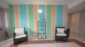 Un pasillo con dos sillones de mimbre con cojines blancos, una estantería metálica y una pared a rayas.