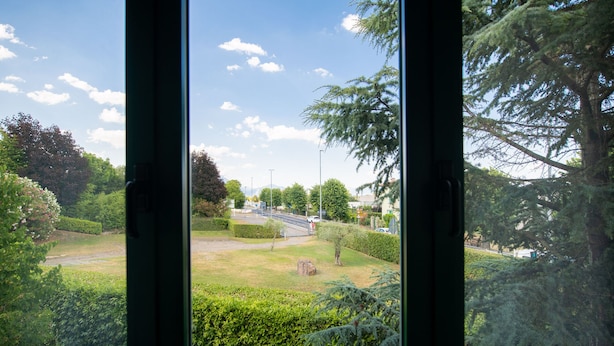 A view through a window of a well-maintained garden with a paved path, trees, and a distant road. - Luxury Martina Appartamenti-2-ortensia (Moniga del Garda)
