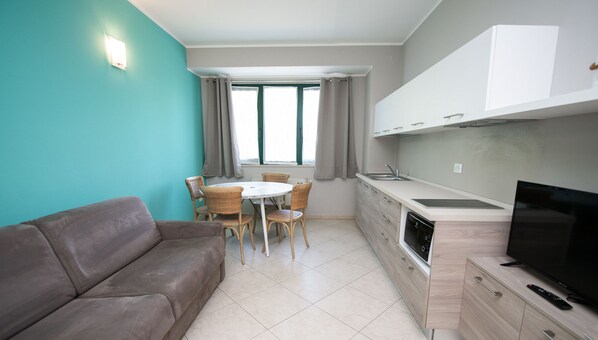 A compact living space with a sofa, dining table, chairs, a kitchen area with a sink and stove, and a TV. - Luxury Martina Appartamenti-2-ortensia (Moniga del Garda)