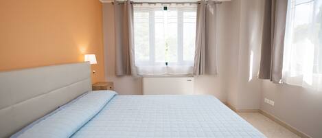 Habitación de hotel con cama, mesita de noche, ventana con cortinas y una lámpara fijada en la pared.