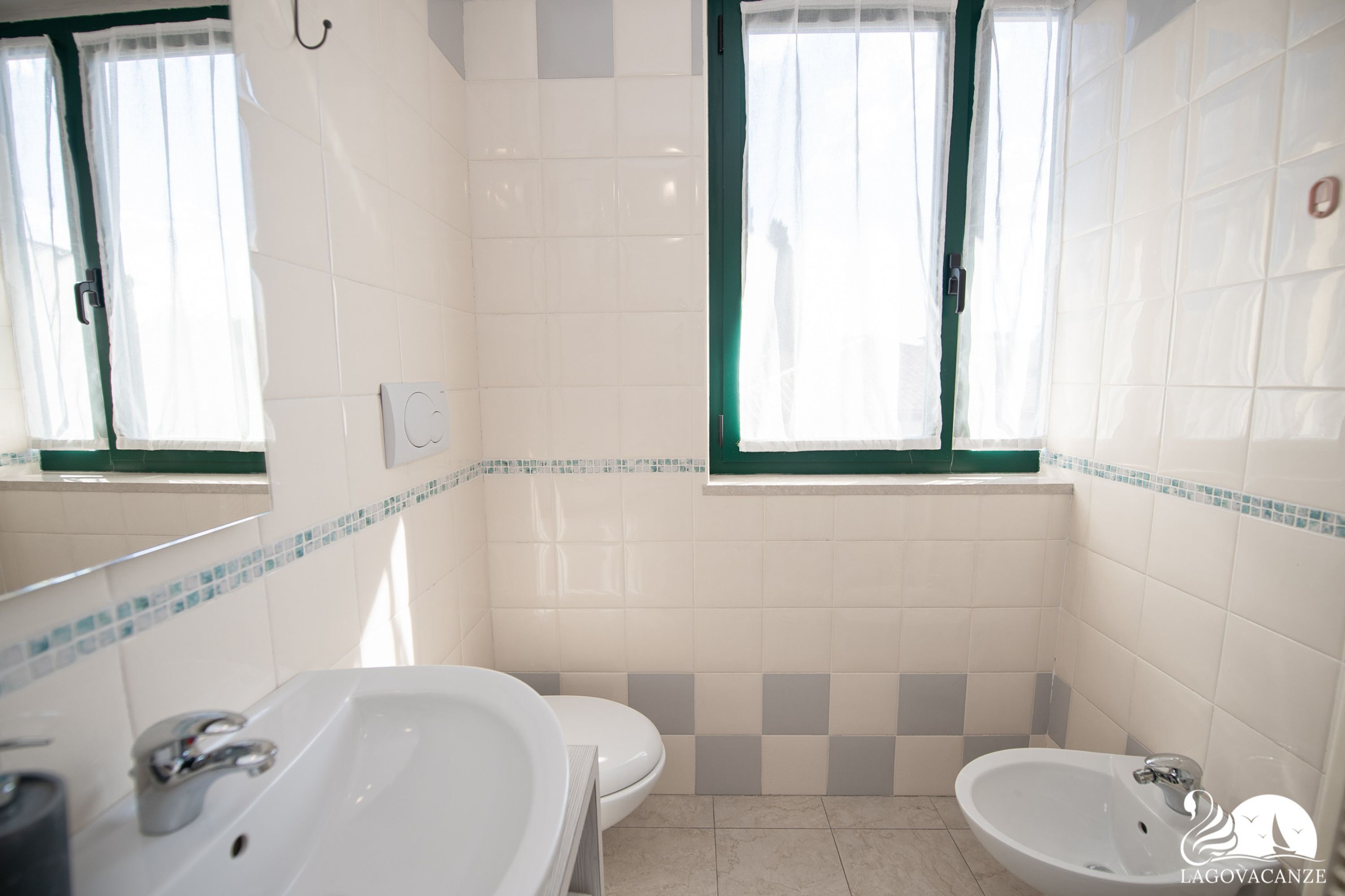 A bathroom with a toilet, a sink, and a bidet. There are two windows with curtains, and the walls are tiled with a checkered pattern.