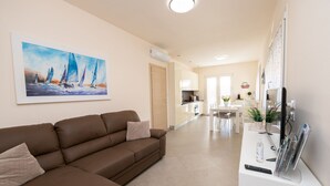 A modern living room with a brown sofa, a painting of sailboats, a kitchen in the background, and a TV stand with a remote control. - Appartamento Lilli in Manerba del Garda (Manerba del Garda)