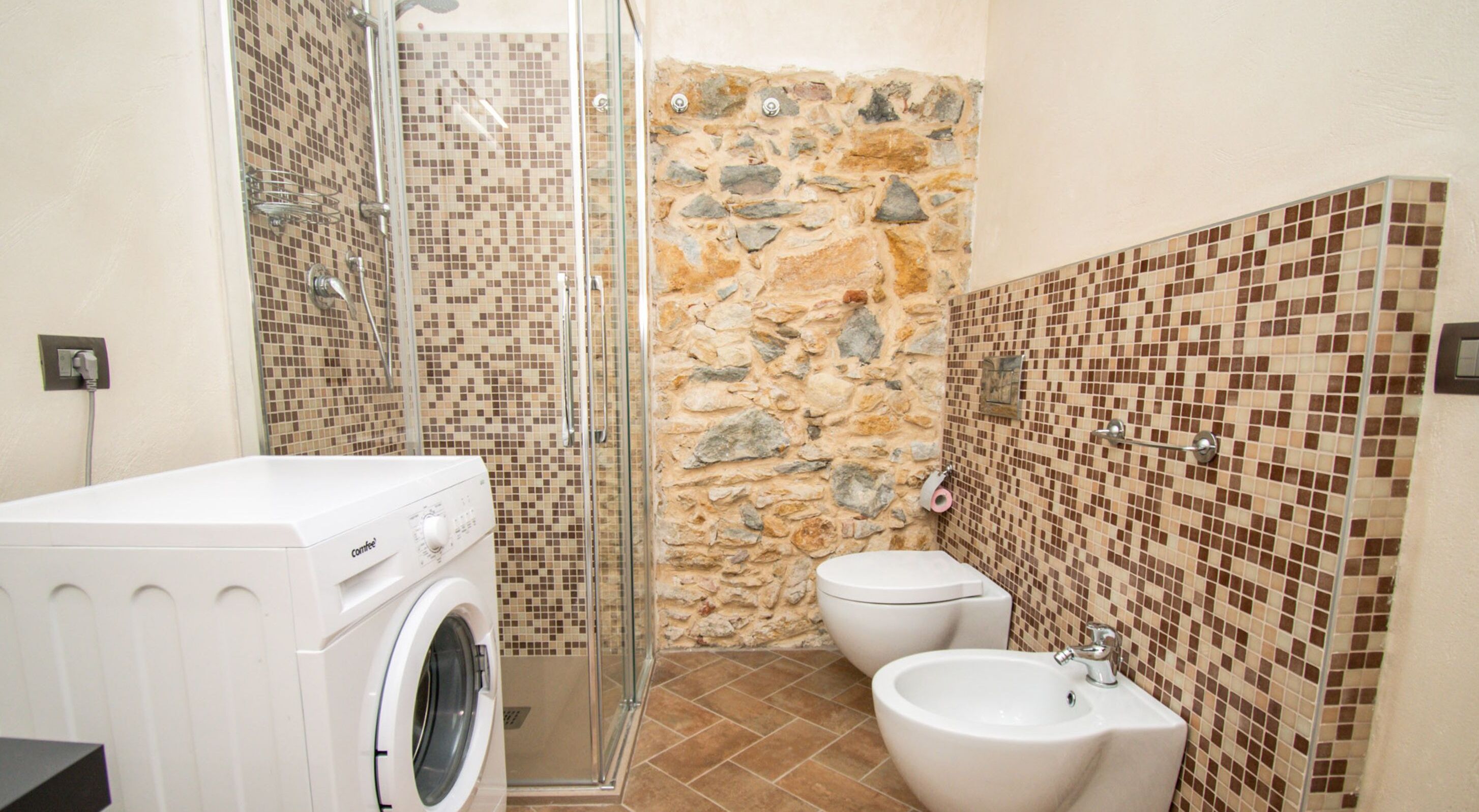 A bathroom with a washing machine, a bidet, and a shower area with mosaic tiles.