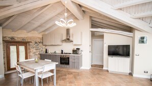 A spacious kitchen with a stone wall, wooden ceiling beams, a dining table with chairs, a built-in oven, a microwave, and a large TV on the wall. - Luxus Gardoncino - La Terrazza sul Lago (Manerba del Garda)
