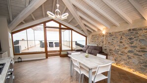 A spacious room with a high ceiling, a stone wall, a large window, and a dining area with a table and chairs. - Luxus Gardoncino - La Terrazza sul Lago (Manerba del Garda)