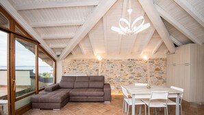 A spacious room with a stone wall, a brown sectional sofa, a dining table with chairs, and a large window offering a view. - Luxus Gardoncino - La Terrazza sul Lago (Manerba del Garda)