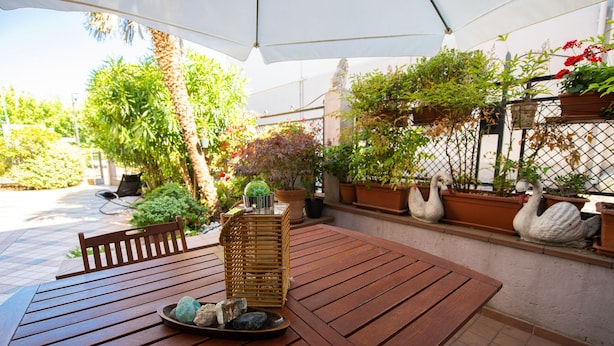 Een houten tafel met een mand en stenen, omringd door potplanten en een zwaanbeeldje onder een witte parasol.