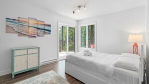 Ein Schlafzimmer mit einem Bett, einem Nachttisch, einer Lampe und einem Fenster mit Blick ins Grüne.