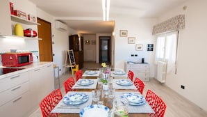 Un comedor con una larga mesa dispuesta para seis personas, rodeada de sillas rojas. Hay un microondas rojo en la encimera a la izquierda, y un estante arriba con diversos objetos.