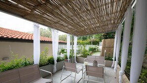 Un patio cubierto con columnas blancas, asientos acolchados y vista a un jardín con techo de tejas rojas al fondo.