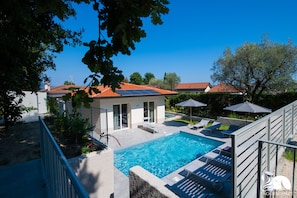 Piscina con sillones de descanso y sombrillas, rodeada por una zona vallada con vistas a casas y árboles.