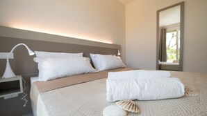 Un lit bien fait, avec une couverture beige, deux oreillers, une serviette blanche et un coquillage.