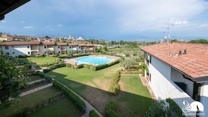 A residential area with a swimming pool surrounded by well-maintained gardens and a view of distant buildings and mountains. - Appartamento Katy in Polpenazze del Garda (Polpenazze del Garda)