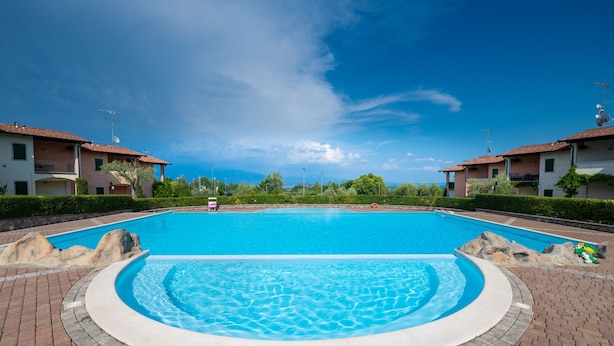 A large outdoor swimming pool surrounded by brick walkways, with two-story buildings and a clear blue sky in the background. - Appartamento Katy in Polpenazze del Garda (Polpenazze del Garda)