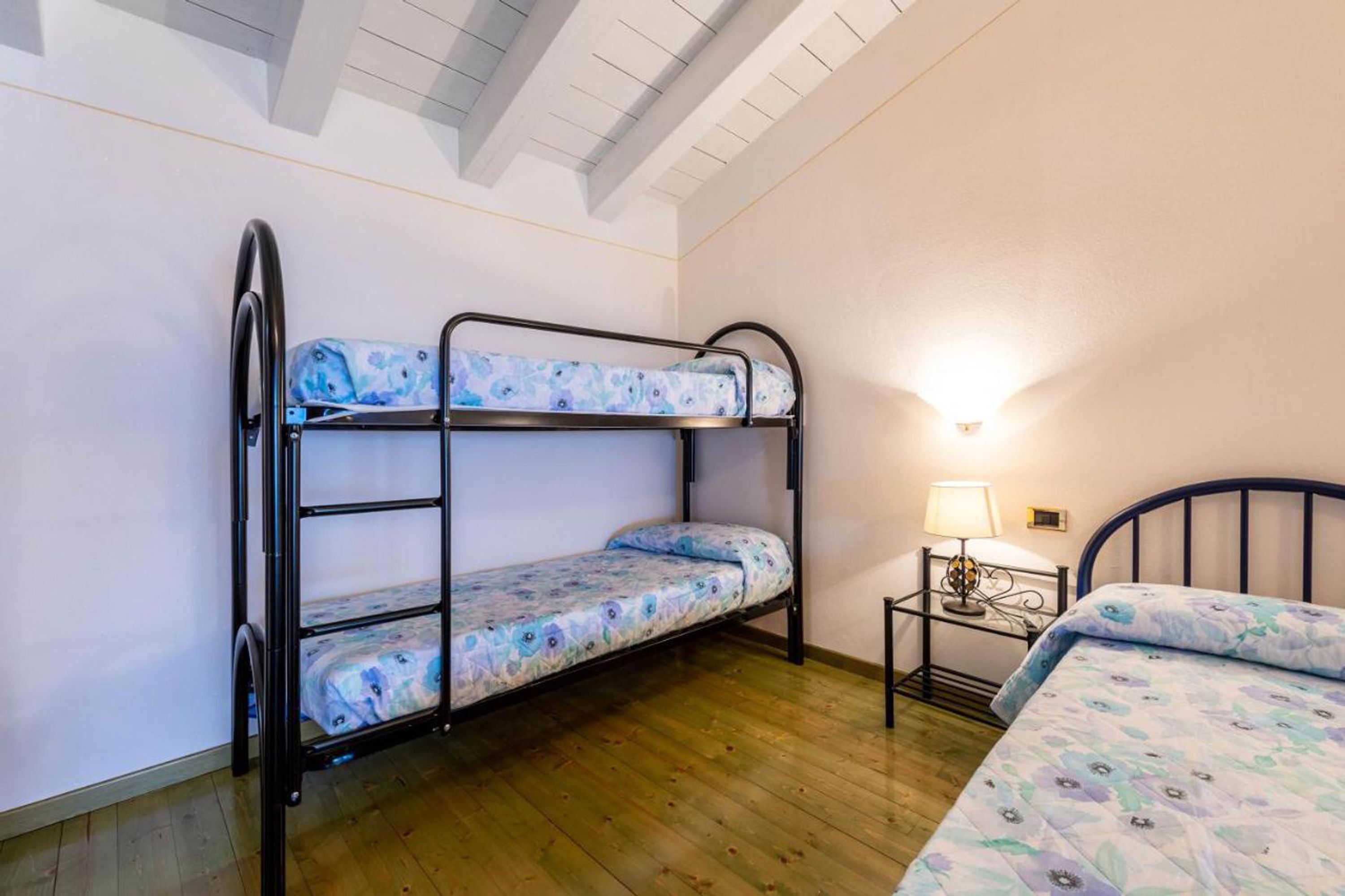 Una stanza con letto a castello, pavimento in legno, un comodino con lampada e un letto singolo con copripiumino a motivi floreali.