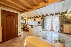 A spacious living area with a wooden ceiling, a stone fireplace, and a kitchen in the background. - Casa Vacanze Baccolo - Appartamento Flavia (Polpenazze del Garda)