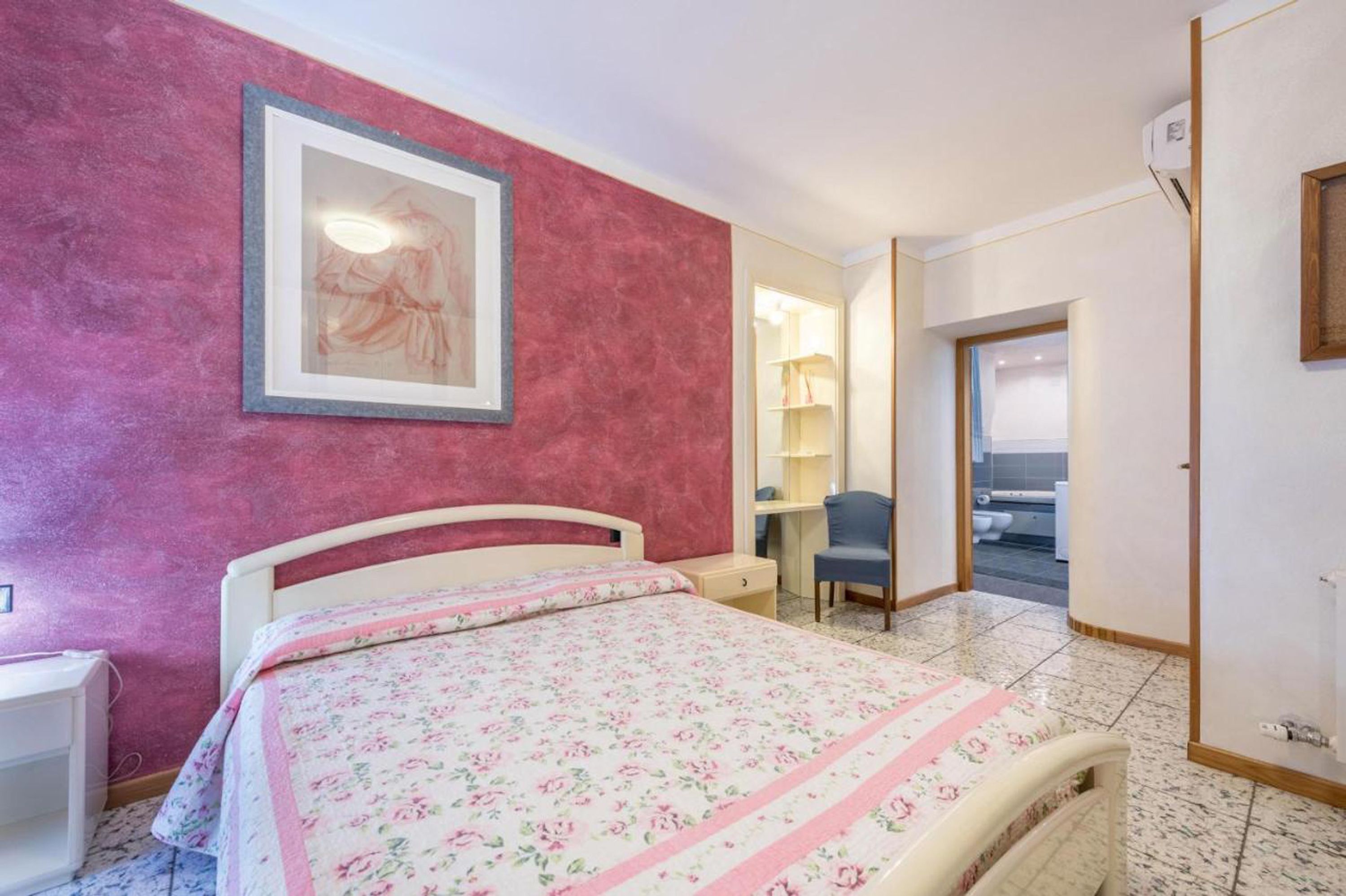 Quarto com cama, escrivaninha, cadeira e banheiro visível por uma porta aberta.