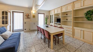 A dining area with a table and chairs, a blue sofa, and a kitchen area with cabinets and a plant. - Casa Vacanze Baccolo - Appartamento Lucia (Polpenazze del Garda)