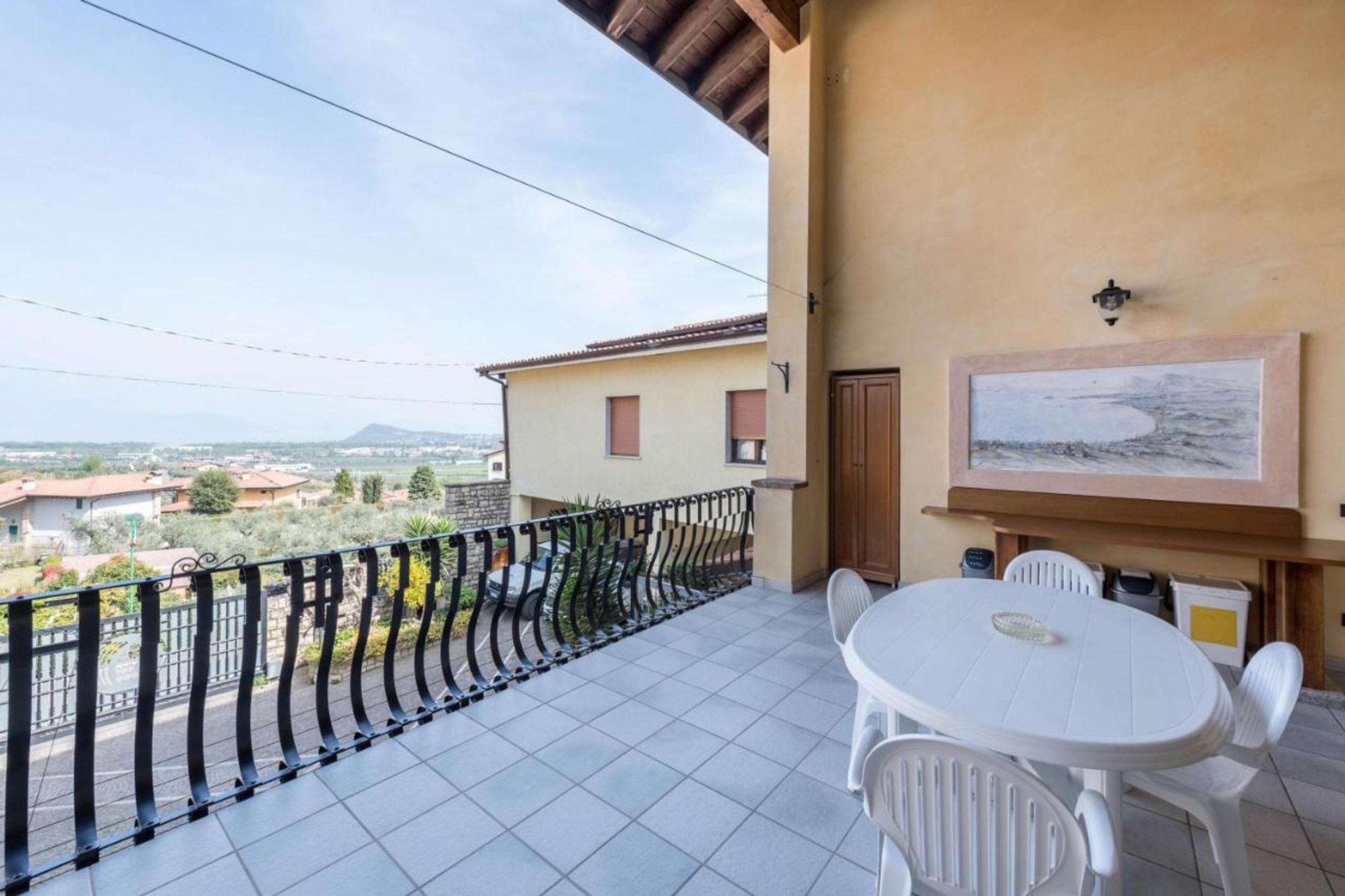 A balcony with a tiled floor, a white table and chairs, a painting on the wall, and a view of houses and mountains in the distance.