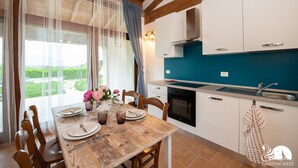Un comedor con mesa de madera dispuesta para cuatro, una cocina con armarios blancos y salpicadero azul, y vista a la vegetación exterior.