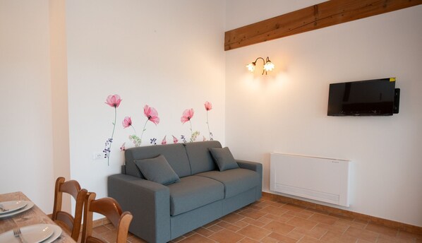 A living room with a grey sofa, wooden chairs, a dining table with plates, a wall-mounted TV, and a radiator. - Il Podere del Bardo - Appartamento Ginevra (Puegnago del Garda)