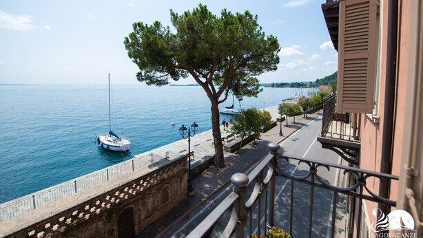 A coastal view with a tree-lined street, a boat docked near the water, and a building with a balcony.