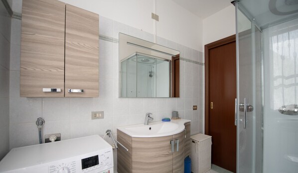 A modern bathroom with a wooden vanity, a large mirror, a glass shower enclosure, and a white sink. - Appartamento Stefano (Polpenazze del Garda)