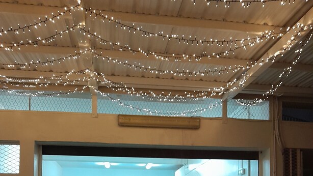 A room with a large, round, dark-colored table in the foreground, a dining area with chairs and a table in the background, and string lights hanging from the ceiling.