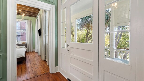 A hallway with wooden flooring, a bed, and a door leading to a balcony with a view of trees. - Beautifully Restored 3-Bedroom Home with Private Ensuite (Charleston)