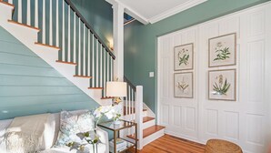 Eine Treppe mit weißen Geländern und Holzstufen, eine weiße Tür mit botanischen Motiven, ein weißes Sofa mit blumigen Kissen und ein kleiner Beistelltisch mit Lampe und Pflanze.