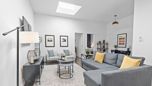 Una sala de estar moderna con sofá, sillón y mesa de centro. Hay cuadros enmarcados en la pared y una lámpara sobre una mesita.
