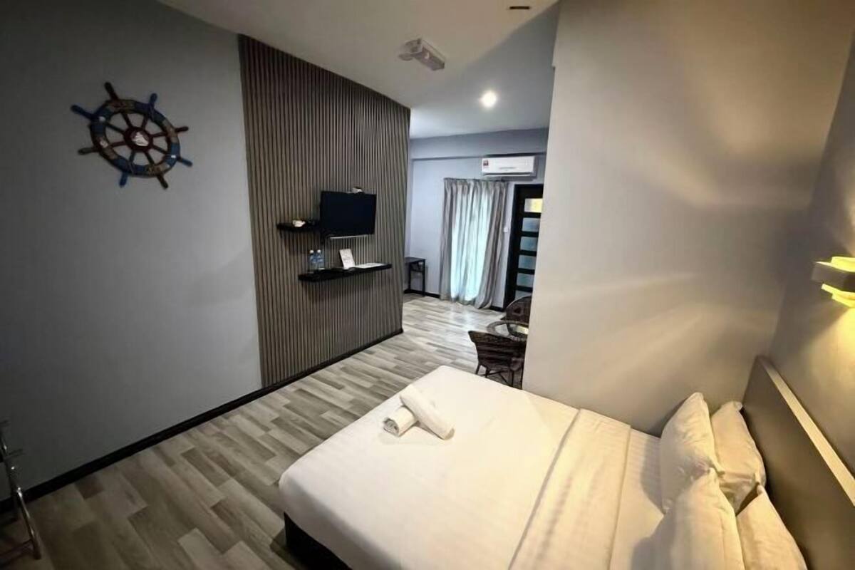 A hotel room with a bed, a TV, a ship's wheel decoration, a wooden panel wall, and a window with curtains.