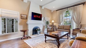 A spacious living room with a fireplace, a television mounted above it, and a large window with curtains. - Spanish Villa Style With Luxury Furniture & Yard! (Los Angeles)