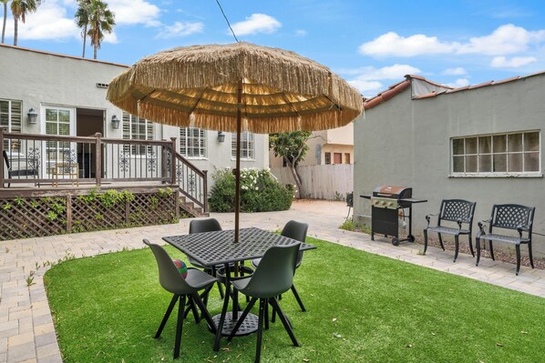 Eine Terrasse mit Tisch, Stühlen, einem Reetdachschirm und einem Grill.
