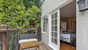 A wooden deck with a wicker chair and table, a bedroom with a bed and nightstand, and a view of lush greenery. - Spanish Villa Style With Luxury Furniture & Yard! (Los Angeles)