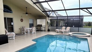 En swimmingpool med liggestole og grillområde, omgivet af en overdækket terrasse.